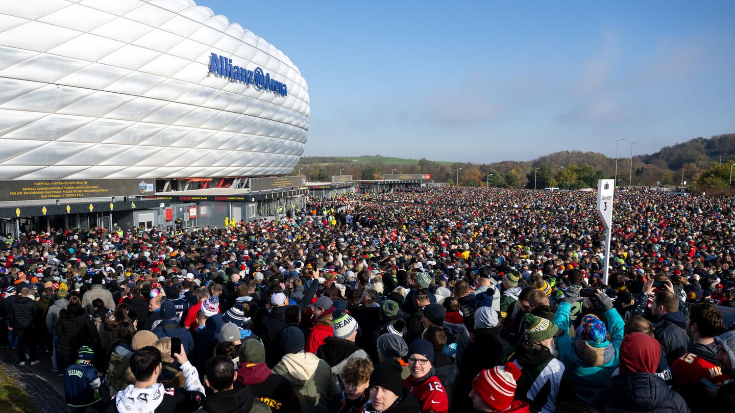 Vor der silbrig-weiß glänzenden Fassade der Allianz-Arena steht eine Menschenmenge Vor der silbrig-weiß glänzenden Fassade der Allianz-Arena steht eine Menschenmenge