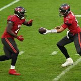 Zwei Männer in rotem Football-Dress laufen aufeinander zu, während einer dem anderen den Ball hinhält