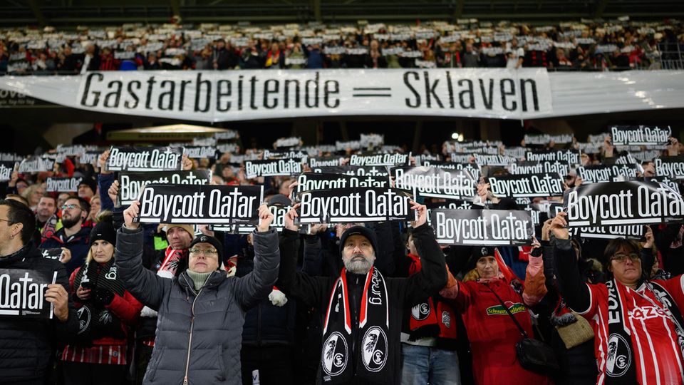 Fans des SC Freiburg stehen mit Plakaten "Boycott Qatar" auf einer Tribüne, an der ein Banner "Gastarbeitende = Sklaven" hängt