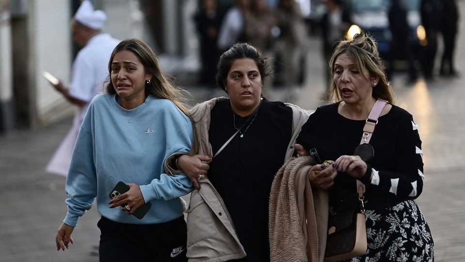 Drei Frauen gehen untergehakt eine breite Straße entlang, während hinter ihnen unscharf Polizei zu erkennen ist