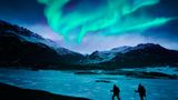 Zwei Wanderer stapfen durch den Schnee in den isländischen Bergen unter dem Polarlicht.