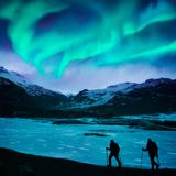 Zwei Wanderer stapfen durch den Schnee in den isländischen Bergen unter dem Polarlicht.