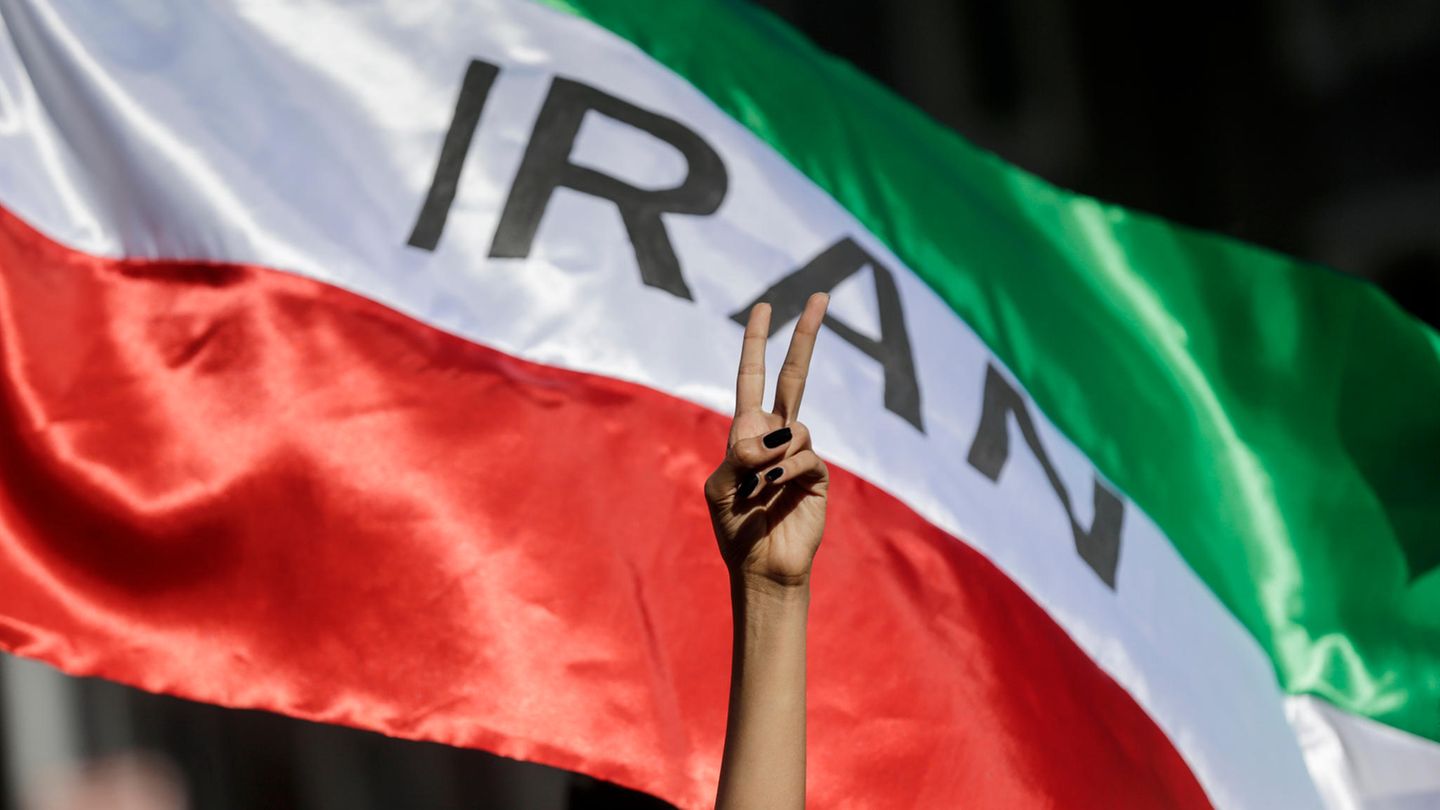 Eine Frau zeigt auf einer Demonstration für die Menschenrechte im Iran das Victory-Zeichen vor einer iranischen Fahne