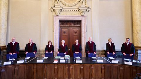 Männer und Frauen in schwarz-roten Roben stehen in einer Reihe hinter einem langen Sitzungstisch unter einem Wappen Berlins