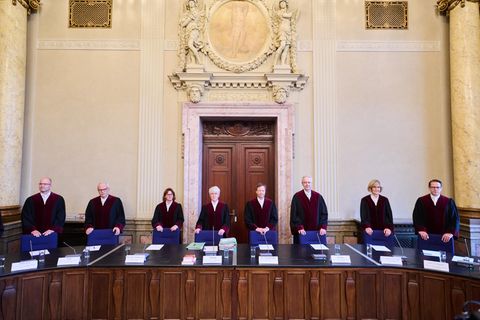 Männer und Frauen in schwarz-roten Roben stehen in einer Reihe hinter einem langen Sitzungstisch unter einem Wappen Berlins