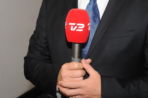 Ein weißer Mann in schwarzem Anzug hält ein Mikrofon mit rotem Windschutz und weißem TV2-Aufdruck in beiden Händen