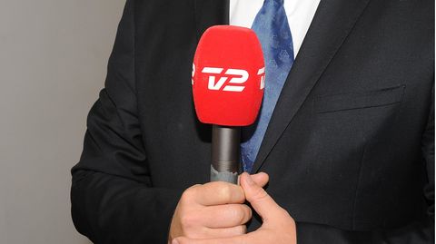 Ein weißer Mann in schwarzem Anzug hält ein Mikrofon mit rotem Windschutz und weißem TV2-Aufdruck in beiden Händen