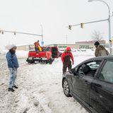 Mehrere Personen stehen auf einer eingeschneiten Straße. Ihr Auto ist offenbar im Schnee stecken geblieben.
