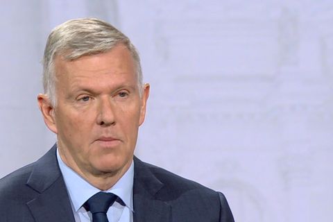 Ein weißer Mann mit weißem Seitenscheitel steht in dunkelblauem Anzug und Krawatte in einem TV-Studio
