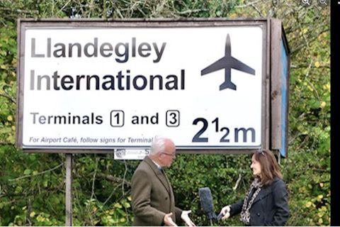 Nicholas Whitehead spricht mit einer BBC-Reporterin vor einem Schild für den fiktiven Llandegley International Airport, Wales
