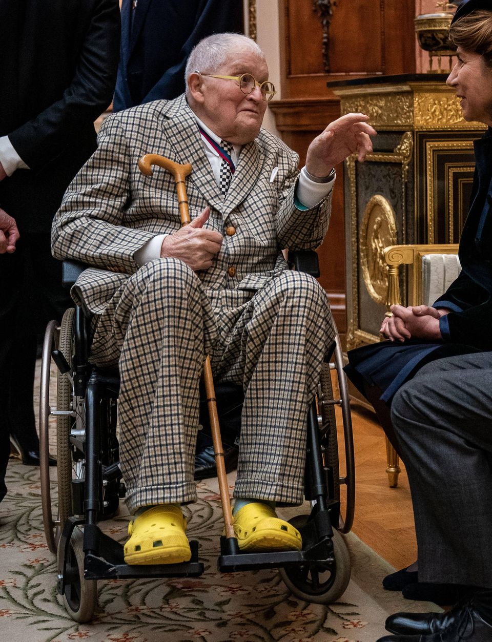 David Hockney sitzt im Rollstuhl und trägt gelbe Plastikschuhe