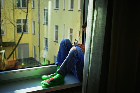 Ein kleiner Junge sitzt alleine hinter einem Vorhang auf der Fensterbank von seinem Zimmer und schaut aus dem Fenster.