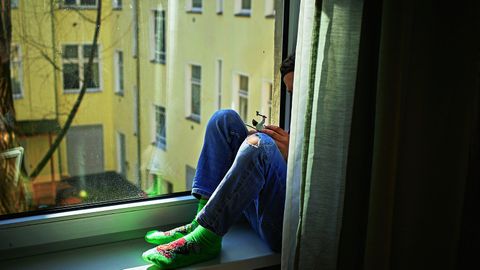 Ein kleiner Junge sitzt alleine hinter einem Vorhang auf der Fensterbank von seinem Zimmer und schaut aus dem Fenster.