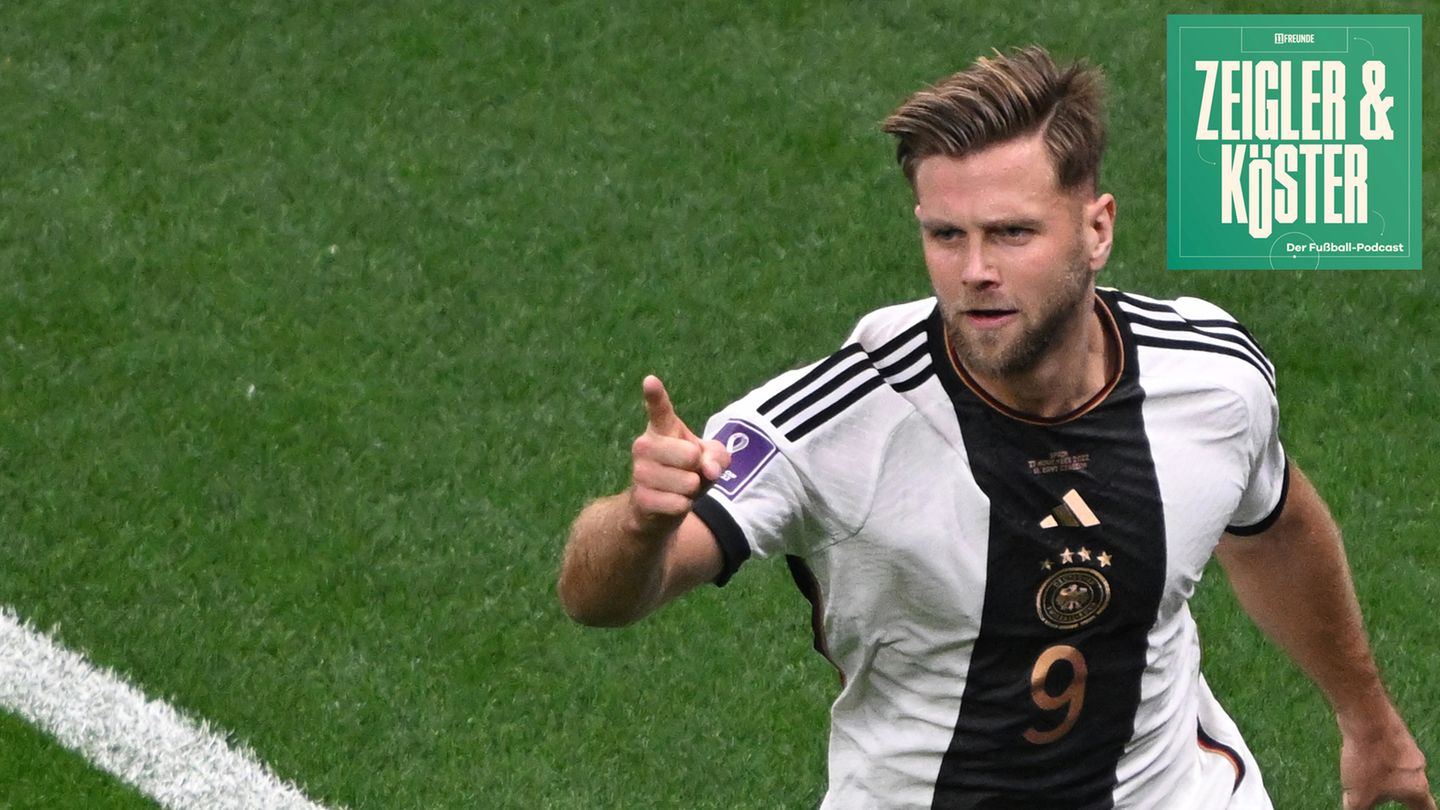 A white professional soccer player in a DFB jersey jubilantly raises his right index finger as he walks across the lawn