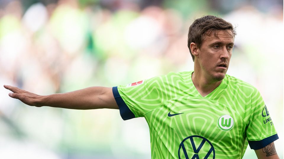Ein weißer junger Man mit Kinn- und Oberlippenbart trägt ein grün-weißes Fußballtrikot mit Wappen des VfL Wolfsburg