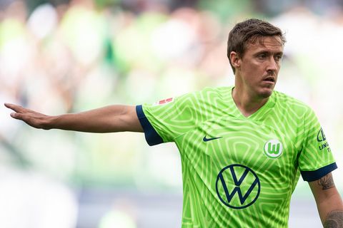 Ein weißer junger Man mit Kinn- und Oberlippenbart trägt ein grün-weißes Fußballtrikot mit Wappen des VfL Wolfsburg