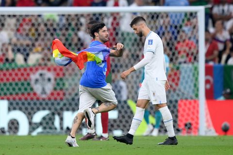 Ein schwarzhaariger Mann mit blauem T-Shirt läuft mit wehender Regenbogenflagge an einem weiß gekleideten Fußballer vorbei