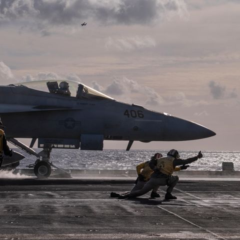 Kampfjet auf der USS George H.W. Bush kurz vor dem Start. Vordergrund geben Leute in der gelber Montur die letzten Befehle.