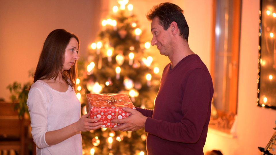 Ein Mann überreicht einer Frau ein Geschenk. Im Hintergrund steht ein Weihnachtsbaum.