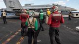 Mehrere Menschen stehen mit verschiedenfarbigen Kitteln auf dem Deck der USS George H.W. Bush, vor ihnen ein Kampfjet