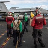 Mehrere Menschen stehen mit verschiedenfarbigen Kitteln auf dem Deck der USS George H.W. Bush, vor ihnen ein Kampfjet