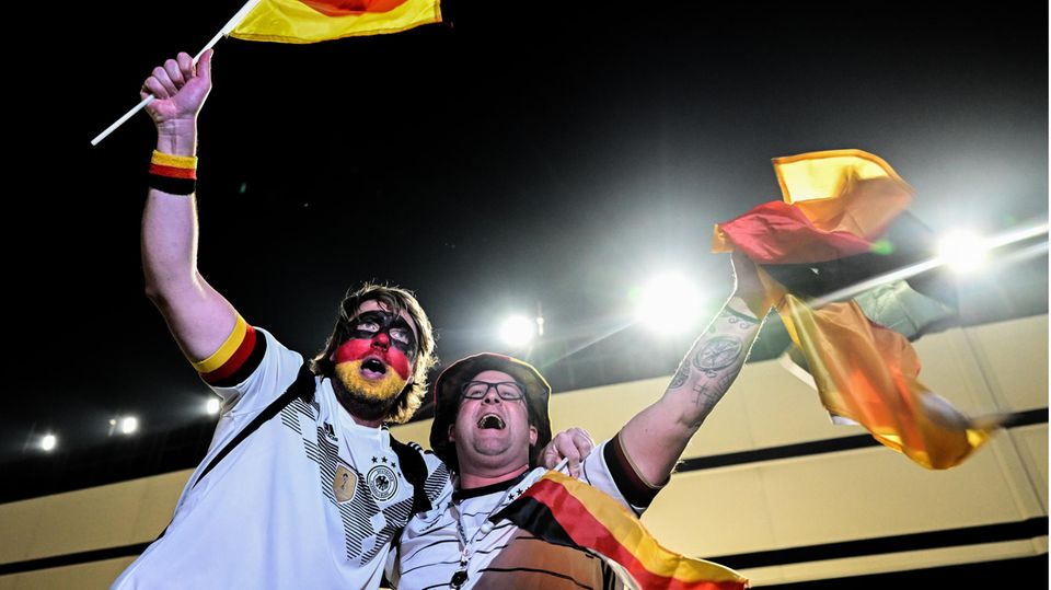 Zwei dickliche weiße Männer in Deutschland-Trikots und mit Flaggen in den Händen jubeln Arm in Arm in einem Stadion