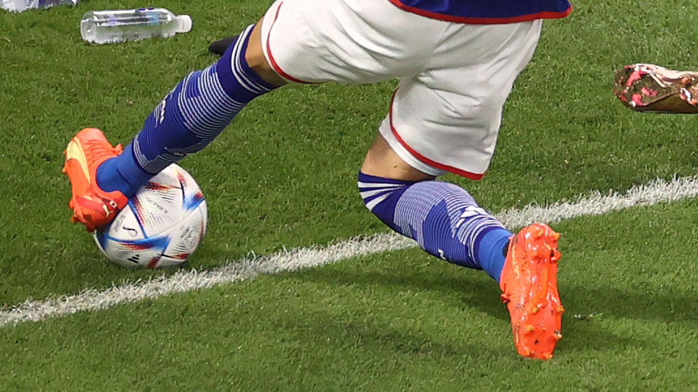 An Asian soccer player in a blue jersey and white shorts plays a ball with his left foot that rolls over the goal line