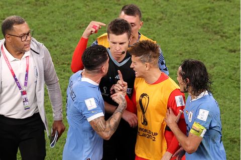Um einen schwarz gekleideten Schiedsrichter hat sich eine Traube aus vier Fußball-Profis und einem Betreuer gebildet