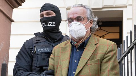 Ein älterer weißer Mann mit zurückgekämmten weißen Haaren wird in Handschellen von zwei vermummten Polizisten abgeführt