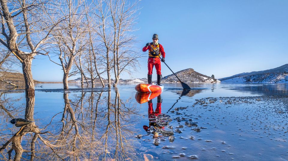 Ein Mann steht in winterlicher Umgebung auf einem SUP-Board auf dem Wasser.
