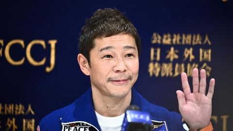 Ein asiatisch aussehender Mann in blauem Oberteil mit Aufnähern winkt mit links ins Publikum