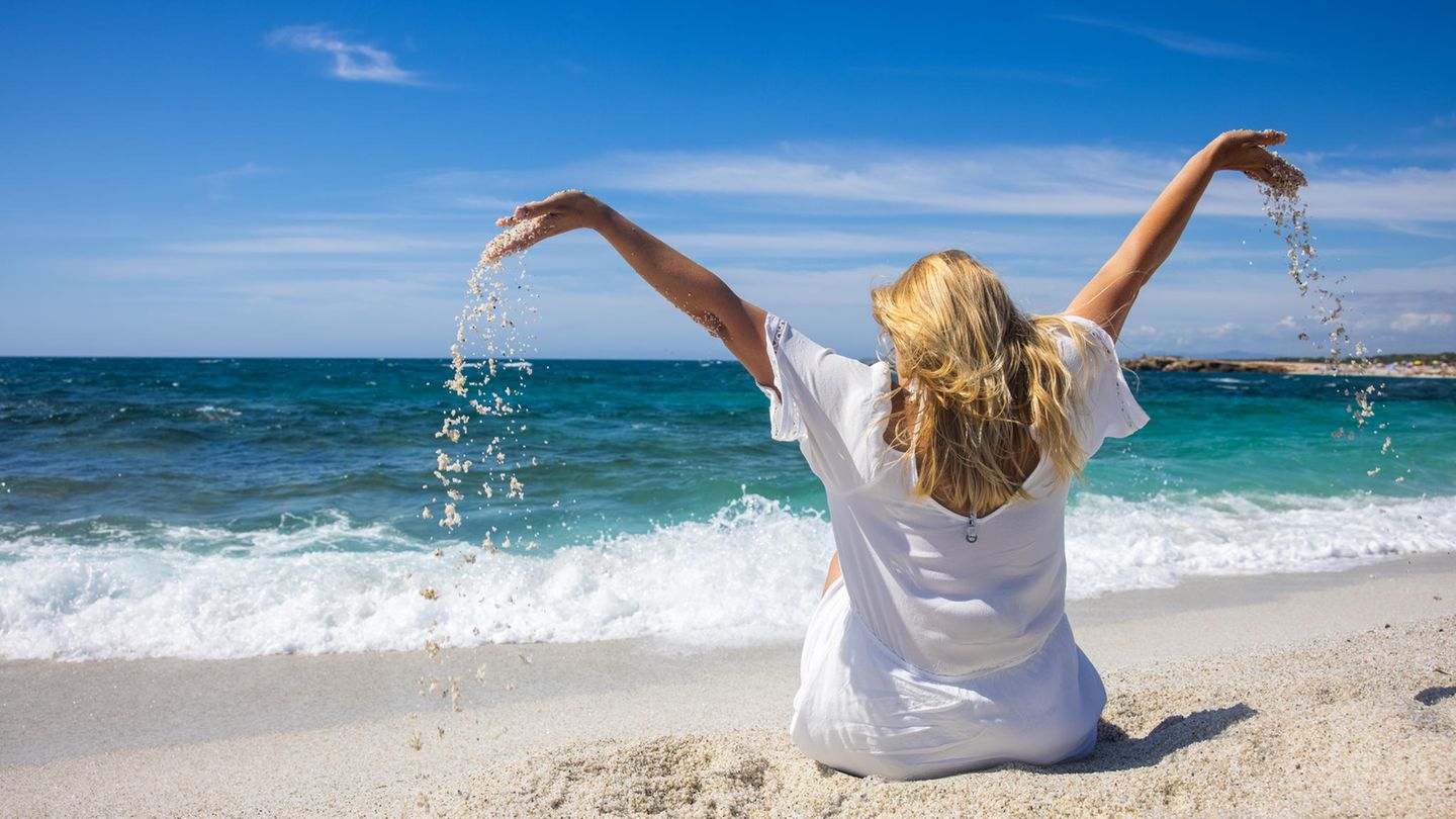 Eine Frau hebt am Strand auf Sardinien mit Sand gefüllte Hände in die Luft.