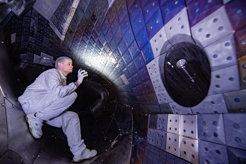 Ein Mann in weißem Overall leuchtet eine gerundete Kachelwand an. Er hockt dabei im Inneren eines Forschungsreaktors
