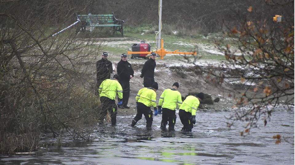 Vier Polizisten in neongelben Jacken gehen durch das knietiefe Wasser im Uferbereich eines Sees. Drei Menschen stehen an Land