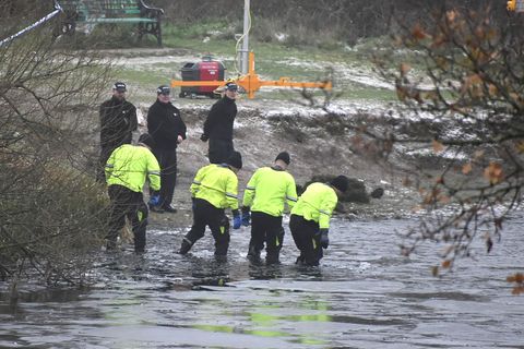 Vier Polizisten in neongelben Jacken gehen durch das knietiefe Wasser im Uferbereich eines Sees. Drei Menschen stehen an Land