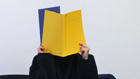 Eine Frau in schwarzem Pulli hält mit beiden Händen einen gelben und einen blauen Pappordner vor ihr Gesicht
