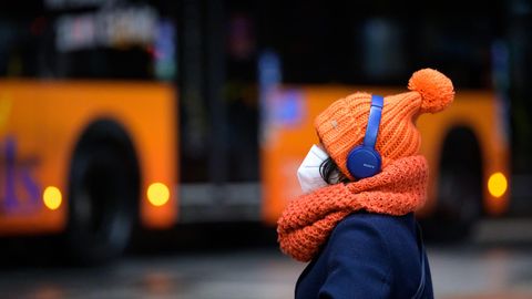 Eine Frau sitzt mit FFP2-Maske, dicker Mütze und Kopfhörern an einem Busbahnhof, während ein Linienbus vorbeifährt