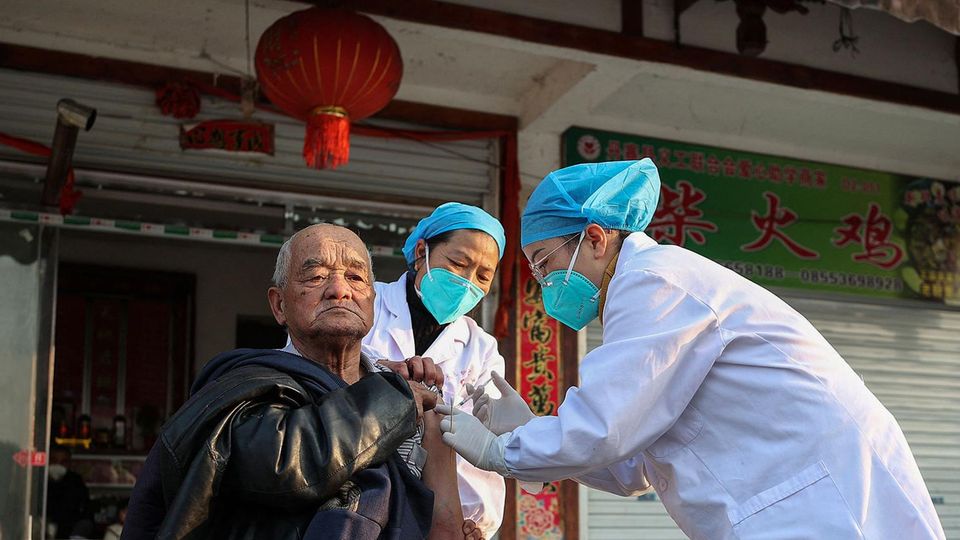 Ein älterer chinesischer Mann bekommt von einer Person mit blauer Haube, grüner Maske und weißem Kittel eine Spritze in den Arm