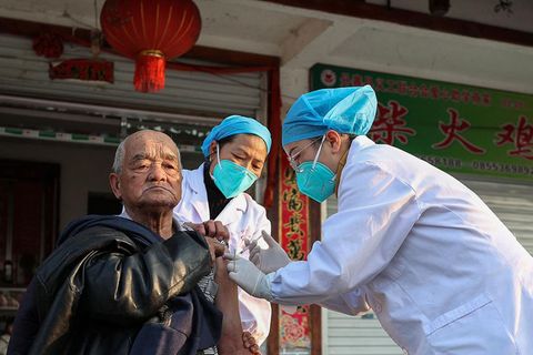 Ein älterer chinesischer Mann bekommt von einer Person mit blauer Haube, grüner Maske und weißem Kittel eine Spritze in den Arm