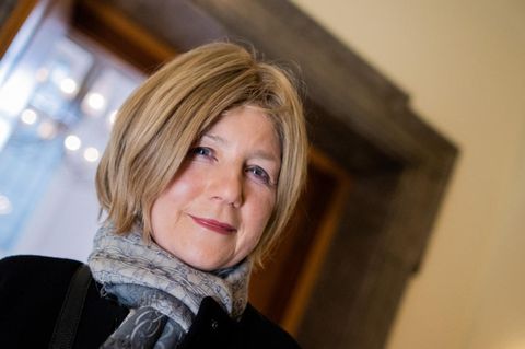 Eine weiße Frau mit blonder Bob-Frisur und blau-grauem Schal lächelt auf dem Flur eines Gerichtsgebäudes