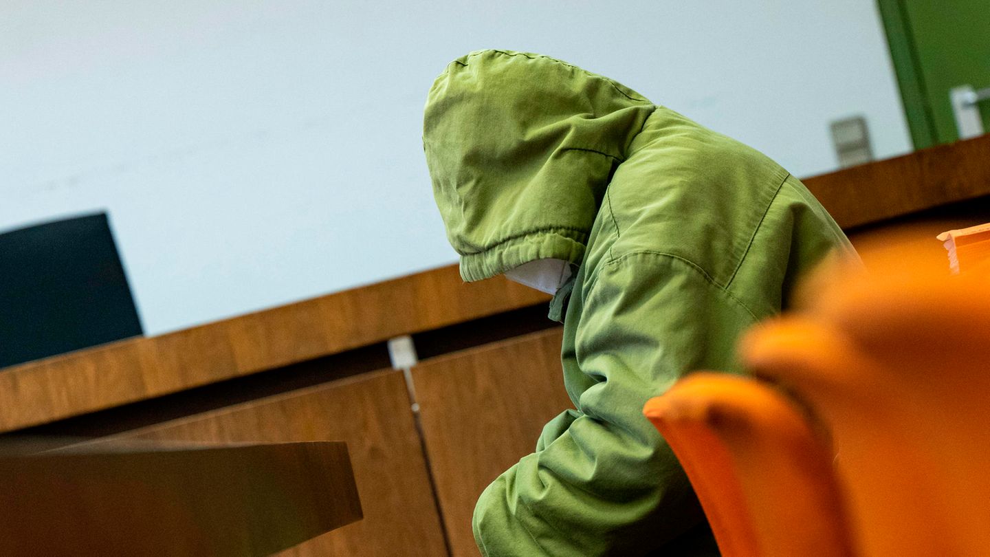 Ein Mann sitzt in einer hellgrünen Jacke mit großer Kapuze auf dem Kopf gebeugt in einem Gerichtssaal