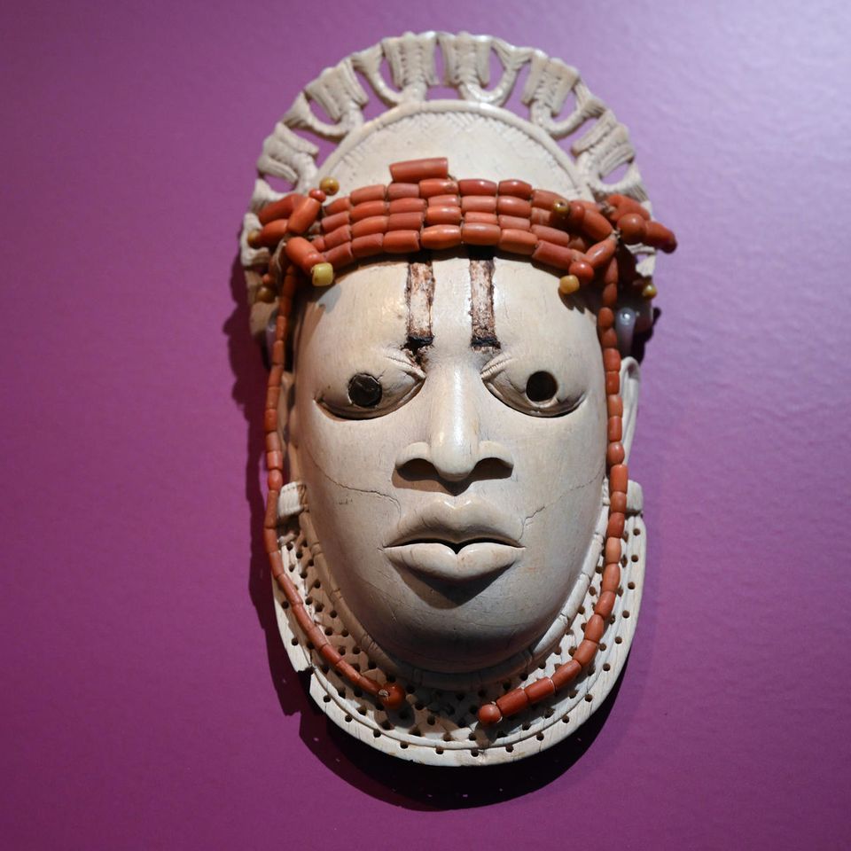 Eines der wertvollsten Stücke der Benin-Bronzen in Deutschland ist diese Miniatur-Elfenbeinmaske. Es gibt nur etwa vier vergleichbare Stücke auf der Welt. Museumsaufzeichnungen zufolge wurde sie 1897 im Schlafgemach des regierenden Königs Ovonramwen gefunden und gestohlen. Es wurde also offensichtlich jahrhundertelang als Ahnenobjekt verehrt und bewacht. Dargestellt ist die Königinmutter ("Iyoba") Idia, die Mutter des 17. Oba Esigie (regierte 1504-1550).