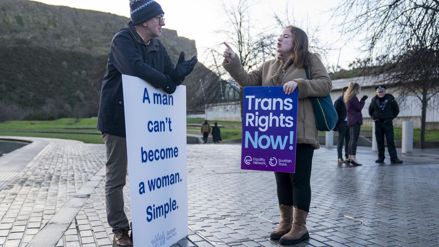 A young woman and an elderly man face each other, both holding protest signs, gesturing