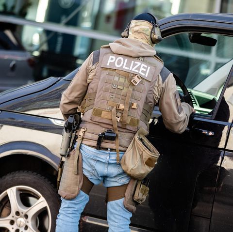 Ein Mann mit Polizei-Schutzweste und Waffen im Holster spricht durch das Fenster eines VW-Buses mit der Person am Steuer