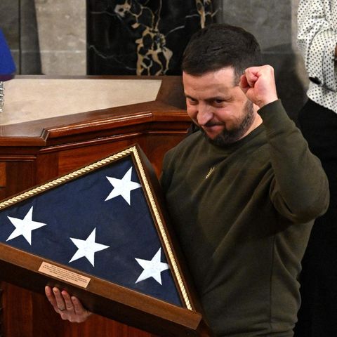 Ein weißer Mann mit Vollbart reckt die linke Faust in die Höhe, während er mit rechte eine eingerahmte US-Flagge hält