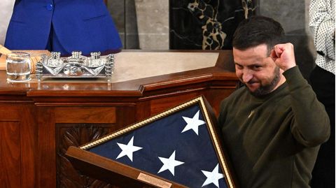 Ein weißer Mann mit Vollbart reckt die linke Faust in die Höhe, während er mit rechte eine eingerahmte US-Flagge hält