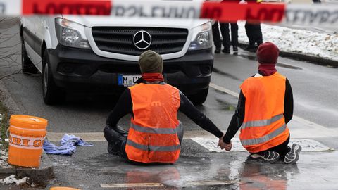 Zwei junge Menschen in orangen Warnwesten knien Hand in Hand in München auf einer Straße