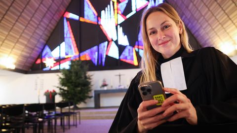 Die junge Pastorin Josephine Teske steht in einer Kirche und hält ihr Smartphone in der Hand