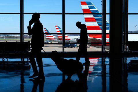Flugreisende gehen an einem Flughafen vor einer Glasfront,  durch die man das Flugfeld mit Flugzeugen sehen kann