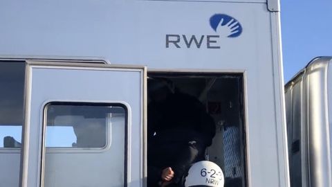 Ein behelmter Polizist schiebt einen Gefesselten die Treppe eines silbernen Gefangenentransporters mit RWE-Logo hoch